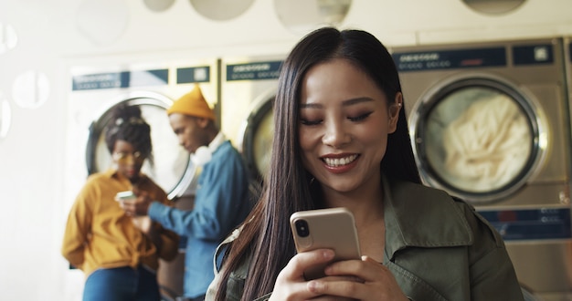 Schließen Sie oben von der asiatischen hübschen fröhlichen Frau, die auf Smartphone im Wäscheservice-Raum tippt und SMS sendet. Schönes glückliches Mädchen, das am Telefon tippt und auf Kleidung wartet, um im Waschhaus sauber zu werden.