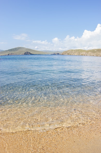 Schließen Sie oben von der sandigen Küste mit einer Welle gegen den Hintergrund des Meeres und der Berge an einem sonnigen Tag