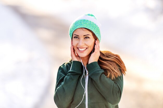 Schließen Sie oben von der schönen kaukasischen Brünette mit Mütze und in der Sportbekleidung, die Musik hört, während Sie sich im Freien ausruhen. Winter Fitness Konzept.