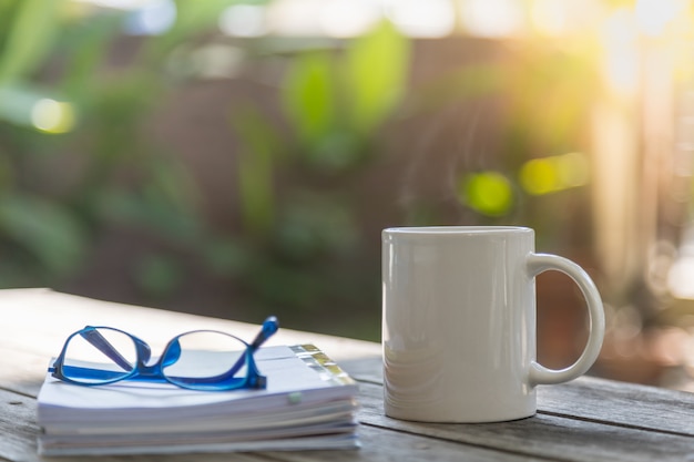 Schließen Sie oben von der weißen Becherschale heißem Kaffee mit Buch und Lesebrille auf Holztisch im Garten mit Kopienraum.