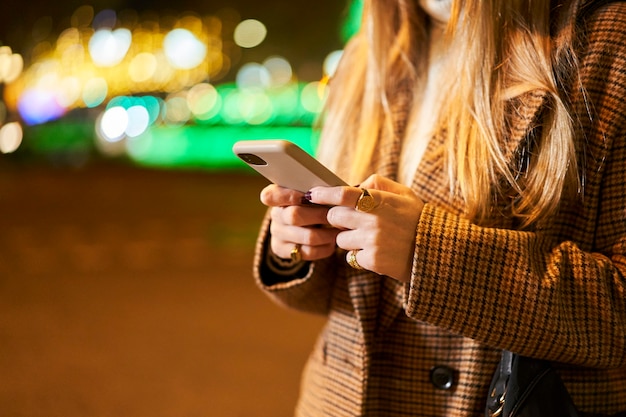 Schließen Sie oben von einer jungen blonden Frau, die ein Smartphone benutzt und eine Nachricht in einer Stadt in der Nacht mit Hintergrundbeleuchtung und Verkehr schreibt.