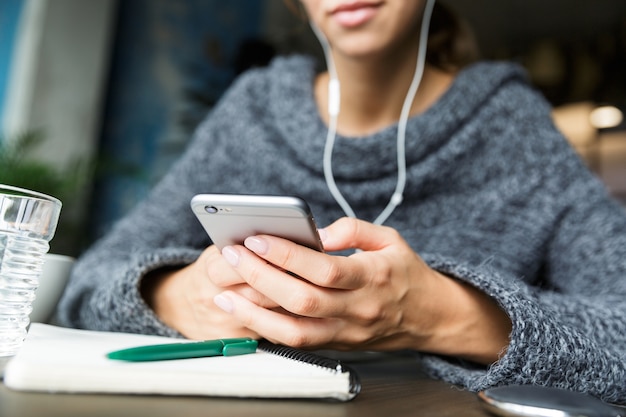 Schließen Sie oben von einer jungen Frau gekleidet im Pullover, der im Stuhl am Kaffeetisch sitzt und Musik mit Kopfhörern hört, unter Verwendung des Mobiltelefons