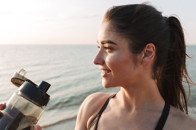 Schließen Sie oben von einer lächelnden jungen Sportlerin, die Wasser trinkt