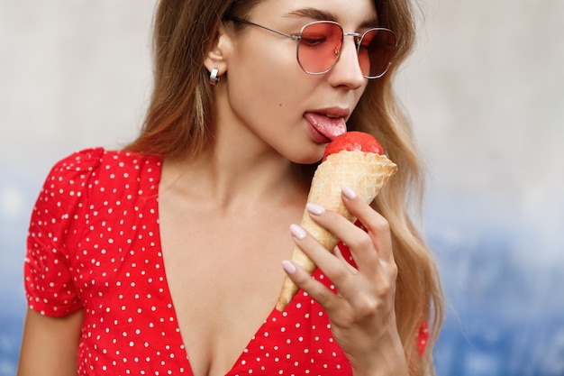 Schließen Sie oben von fasionable blondem Mädchen, das Eis nahe Straßenmauer leckt