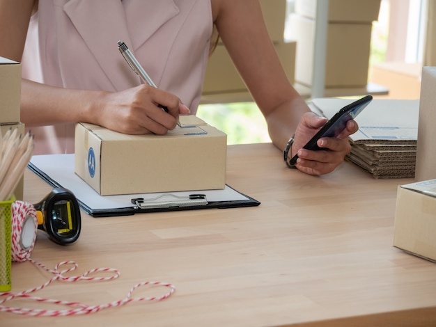 Schließen Sie oben von Frau Handverkäufer, der Adresse auf Paketbox schreibt und Produktbestellung auf Smartphone überprüft