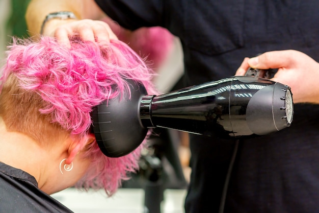 Foto schließen sie oben von friseur, der kurzes rosa oder rotes haar mit einem haartrockner in einem schönheitssalon trocknet