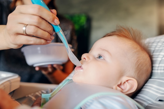 Schließen Sie oben von hungrigem kaukasischem Baby mit Lätzchen, das in seinem Stuhl sitzt und Babynahrung isst. Seine Mutter gab ihm zum ersten Mal Essen.