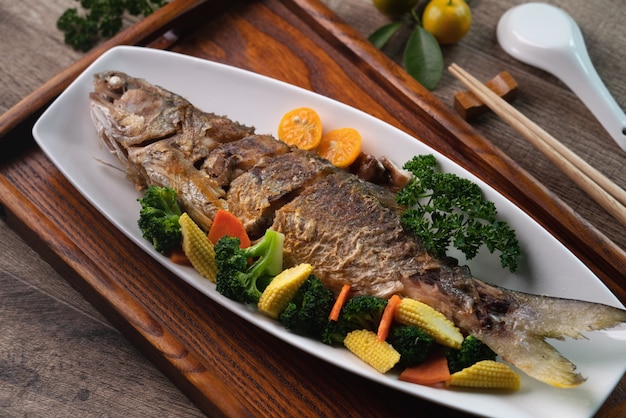 Schließen Sie oben von köstlichem gebratenem Fischgericht in einem weißen Teller mit Gemüse auf einem Holztisch.