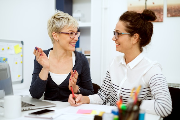 Schließen Sie oben von zwei lächelnden stilvollen Geschäftsfrauen mittleren Alters, die arbeiten und ein Gespräch haben, während sie im Büro nebeneinander sitzen.