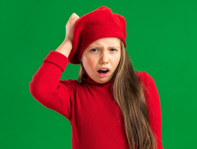 Schmerzendes kleines blondes Mädchen mit rotem Barett, das nach vorne schaut und die Hand auf dem Kopf isoliert auf grüner Wand hält