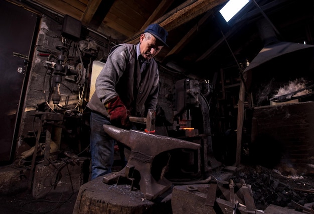 Schmied, der das rotglühende geschmolzene Metall auf dem Amboss in der traditionellen Schmiedewerkstatt manuell schmiedet. Schmied, der Metall mit Hammer in der Schmiede bearbeitet