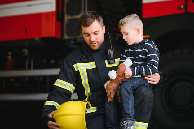 Schmutziger Feuerwehrmann in Uniform mit kleinem geretteten Jungen, der auf schwarzem Hintergrund steht