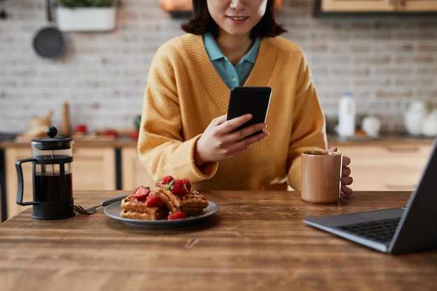 Schnappschuss einer lächelnden asiatischen Frau, die ein Smartphone benutzt, während sie das Frühstück in einem gemütlichen Kopierraum in der Küche zu Hause genießt
