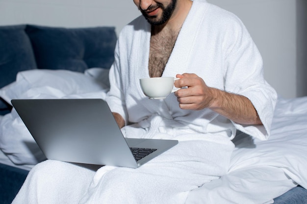 Schnappschuss eines bärtigen jungen Mannes, der einen Laptop benutzt, während er im Bett Kaffee trinkt