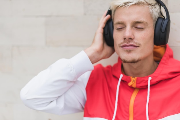 Schnappschuss eines gutaussehenden Mannes mit blonden Haaren in roter Sportbekleidung, der sich nach körperlichen Aktivitäten an der grauen Wand draußen entspannt und sitzt Stilvoller Mann, der Musik über Bluetooth-Kopfhörer hört