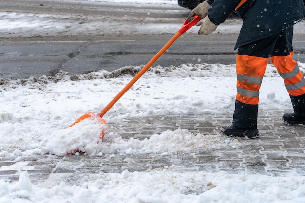 Schnee vom Bürgersteig nach Schneesturm entfernen Ein Straßenarbeiter mit einer Schaufel in den Händen und in Spezialkleidung reinigt den Bürgersteig und die Straße vom Schnee Schneesturm und Orkan in der Stadt