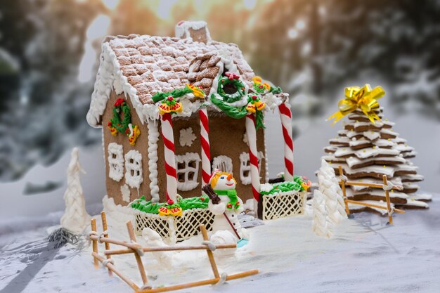 Schneebedecktes hausgemachtes Lebkuchenhaus, Lebkuchen-Weihnachtsbaum und ein Zuckermastix-Schneemann auf defokussiertem Waldhintergrund. Mockup für saisonale Angebote und Urlaubspostkarte