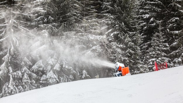 Schneekanone sprüht künstliche Eiskristalle auf die Skipiste, Beschneiung im Wintersportort, Bäume im Hintergrund