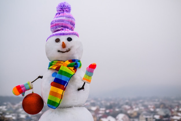 Schneemann hält Ballschneemann des neuen Jahres im Winterhutweihnachtshintergrund mit lustigem Schneemann des Schneemanns