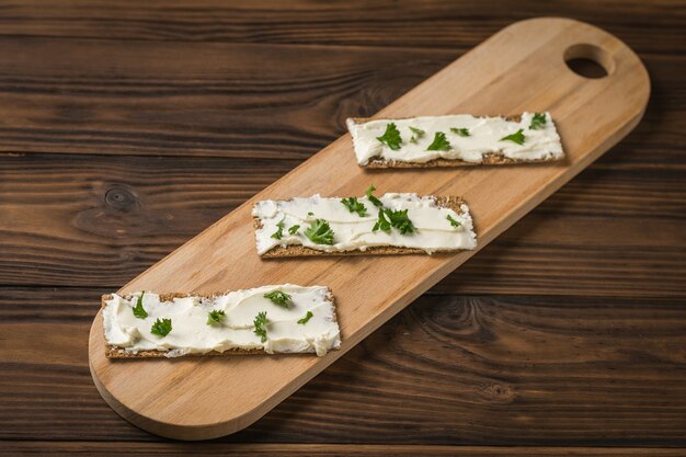 Schneidebrett mit drei Toasts mit Frischkäse und Kräutern auf einem Holztisch. Vegetarischer Snack. Gemüse mit Frischkäse und Brot.