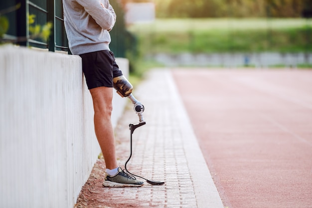 Schneiden Sie das Bild eines gutaussehenden kaukasischen sportlich behinderten Mannes in Sportbekleidung und mit künstlichem Bein aus, das sich auf den Zaun stützt, während Sie die Arme verschränkt halten.