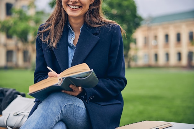 Schneiden Sie das Foto einer Dame mit langen Haaren ab, die draußen sitzt und lächelt, während sie ein Notizbuch mit Bleistift hält holding