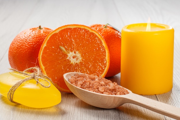 Schneiden Sie Orange mit zwei ganzen Orangen, Seife, Holzlöffel mit gelbem Meersalz und brennender Kerze auf Holzschreibtisch. Spa-Produkte und Zubehör