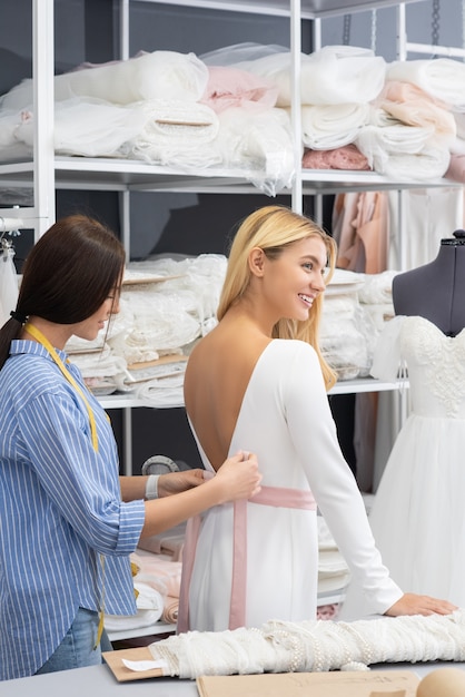 Schneider, der rosa Satingürtel um Taille der glücklichen Braut während des Hochzeitskleides zusammenbindet, das im Schneiderstudio passt