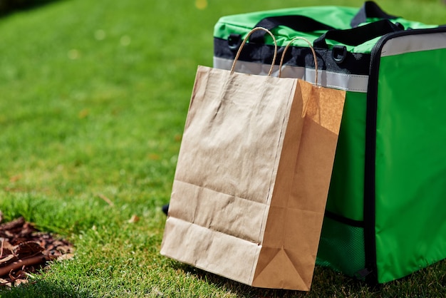 Schnelle Lieferung grüner Thermorucksack und Papiertüte, die auf dem grünen Gras im Freien stehen, nah