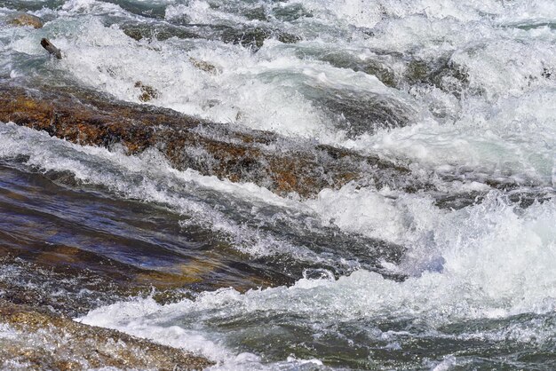 Schneller Frühlingsfluss, der an einem sonnigen Tag über Felsen fließt und Wildwasserwellen bildet, Detail in der Nähe - abstrakter Naturhintergrund