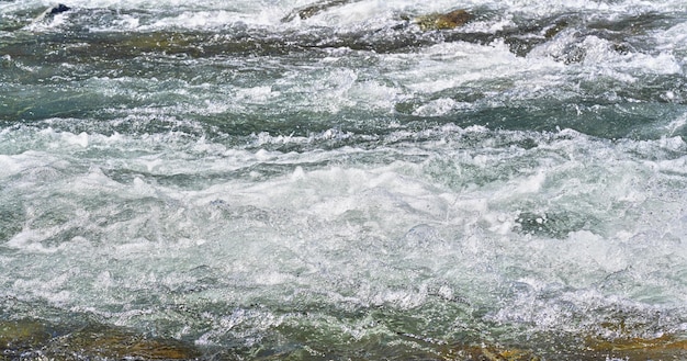 Schneller Frühlingsfluss, der an einem sonnigen Tag über Felsen fließt und Wildwasserwellen bildet, Detail in der Nähe - abstrakter Naturhintergrund.