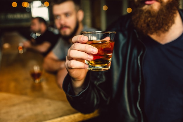 Schnittansicht des berded Mannes in der schwarzen Lederjacke, die Glas mit Alkohol hält. Zwei andere Männer sitzen dahinter. Sie sind konzentriert.