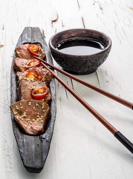 Foto schnitte orientalisches scharfes fleisch auf einem langen dunklen holzteller mit stöcken und einer schüssel mit soße auf der weißen oberfläche mit zerrissener farbe