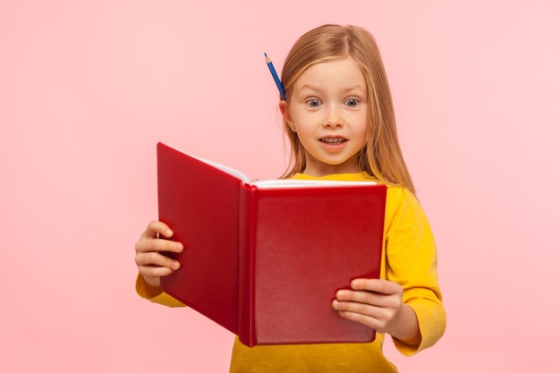 Schockierende Fakten. Erstauntes, intelligentes kleines Mädchen mit Bleistift hinter dem Ohr, das ein großes Buch mit überraschtem Ausdruck liest, erstaunt über die Geschichte, Lernenzyklopädie. Studioaufnahme im Innenbereich isoliert auf rosa Hintergrund