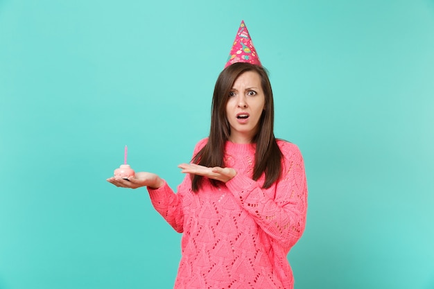 Schockiert betroffene junge Frau in gestricktem rosa Pullover und Geburtstagshut, die Hand auf Kuchen mit Kerze zeigt, die auf blauem Wandhintergrund Studioporträt isoliert ist. Menschen Lifestyle-Konzept. Kopieren Sie Platz.