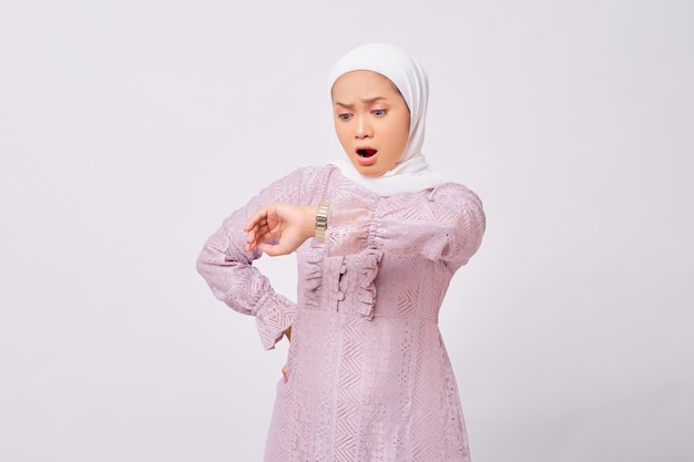 Schockiert schöne junge asiatische Muslimin mit Hijab und lila Kleid hob den Arm und sah Armbanduhr isoliert auf weißem Studiohintergrund aus