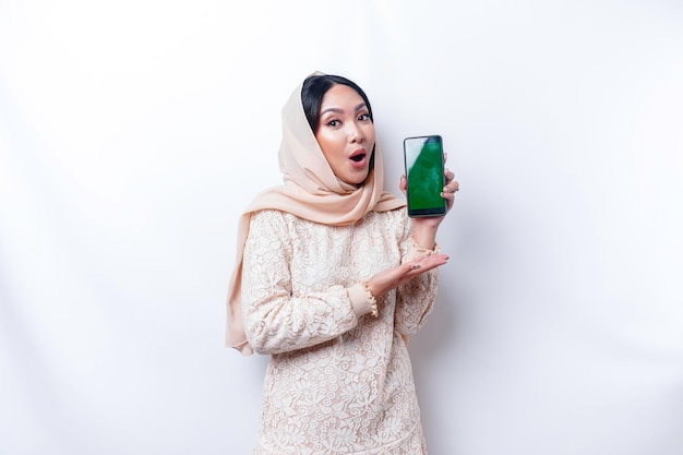 Schockierte asiatische Frau, die Hijab trägt und Kopienraum auf ihrem Telefonbildschirm zeigt, isoliert durch weißen Hintergrund