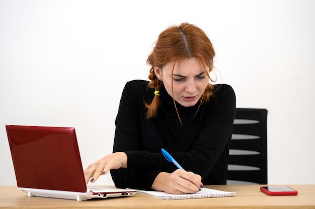 Schockierte ernsthafte junge Büroangestellte Frau, die hinter Schreibtisch mit Laptop-Computer, Handy und Notizbuch sitzt.