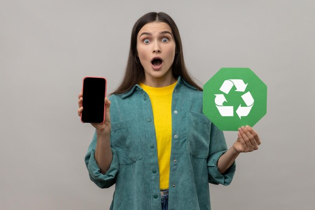 Schockierte erstaunte Frau, die Handy mit leerem Bildschirm und Recycling-Symbol zeigt und in die Kamera blickt