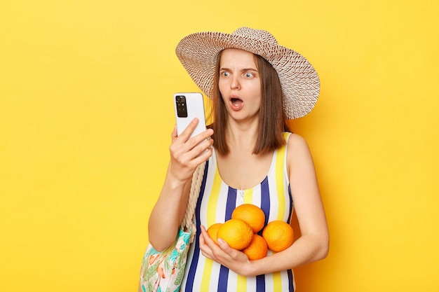 Schockierte, erstaunte Frau in gestreiftem Badeanzug und Hut isoliert auf gelbem Hintergrund, die mit überraschtem Gesichtsausdruck auf ihr Mobiltelefon blickt und den Mund offen hält