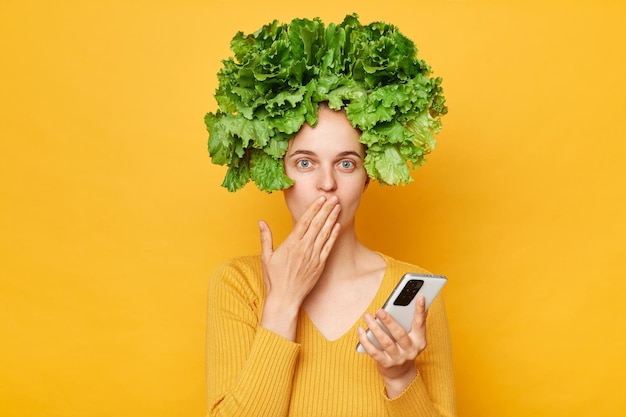 Schockierte Frau in lässigem Hemd und Salatkranz steht isoliert vor gelbem Hintergrund und nutzt ihr Mobiltelefon, um im Internet zu surfen und den Mund mit der Hand zu bedecken