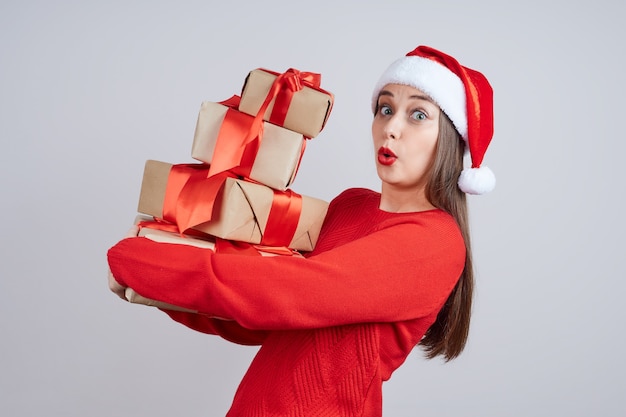 Schockierte Frau in Weihnachtsmütze, roter Pullover, hält ein Bündel Geschenke mit einem roten Band. Feiertagskonzept, Weihnachten, Neujahr.