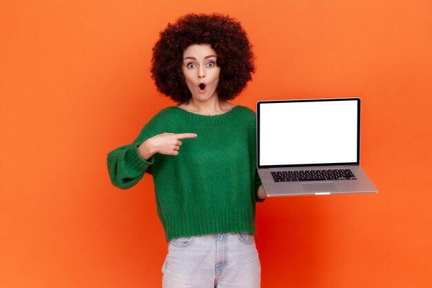 Schockierte Frau mit Afro-Frisur, die einen grünen, lässigen Pullover trägt und auf ein weißes, leeres Display ihres tragbaren Computers zeigt, Kopierraum. Studioaufnahme im Innenbereich isoliert auf orangefarbenem Hintergrund.