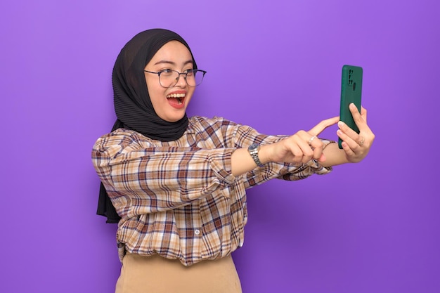 Schockierte junge asiatische Frau in kariertem Hemd, die ein Mobiltelefon benutzt, um großartige Nachrichten isoliert auf violettem Hintergrund zu erhalten