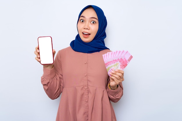 Schockierte junge asiatische muslimische Frau in rosafarbenem Hemd, die ein Handy mit leerem Bildschirm zeigt und Geldbanknoten isoliert über weißem Hintergrund hält Religiöser Lebensstil der Menschen