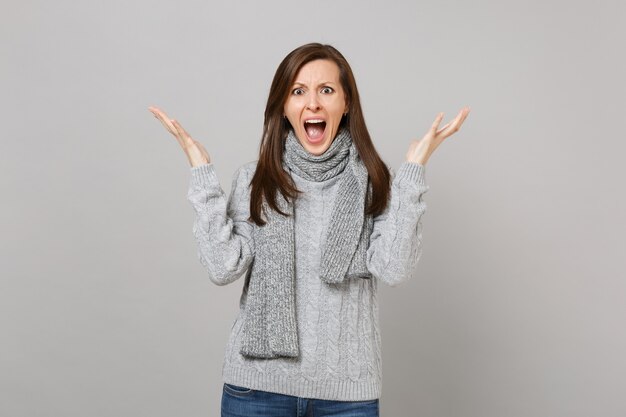 Schockierte junge Frau im grauen Pullover-Schal schreien, die Hände einzeln auf grauem Hintergrund im Studio ausbreitend. Gesunder Modelebensstil, aufrichtige Emotionen der Menschen, Konzept der kalten Jahreszeit. Kopieren Sie Platz.