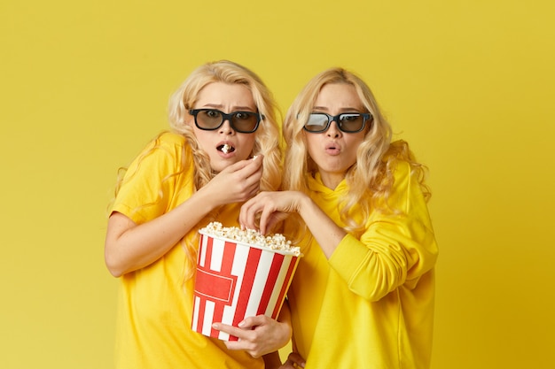 Schockierte junge Models Frauen in 3D-Gläsern essen Popcorn, sieht gruselig aus. Auf gelber Wand isoliert