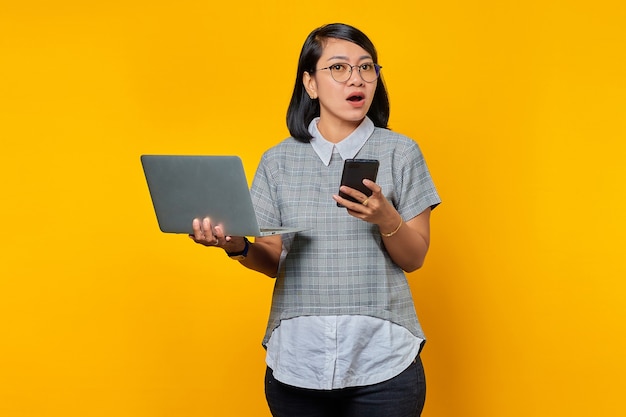 Schockierte junge schöne Frau, die Smartphone und Laptop hält und auf gelbem Hintergrund beiseite schaut