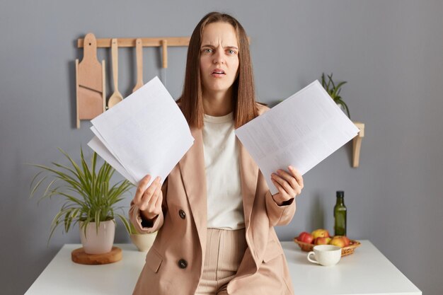 Schockierte kaukasische Frau, die mit Papier arbeitet und Dokumente in Händen hält, die mit unzufriedenem Ausdruck in formelle Kleidung in die Kamera blickt