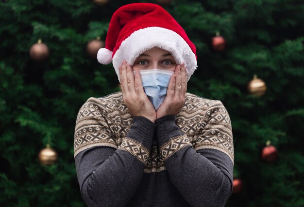 Schockierte oder überraschte Weihnachten. Schließen Sie herauf Porträt des Mannes, der einen Weihnachtsmannhut, Weihnachtspullover und medizinische Maske mit Emotion trägt. Vor dem Hintergrund eines Weihnachtsbaumes. Coronavirus Pandemie